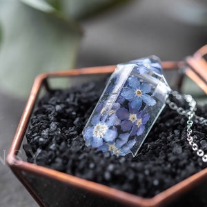 May include: A silver chain necklace with a clear glass pendant containing pressed blue forget-me-not flowers. The necklace is displayed in a hexagonal glass box with a black sand base.