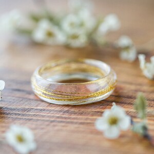 May include: A clear resin ring with gold wire embedded inside. The ring is sitting on a wooden surface with white flowers scattered around it.
