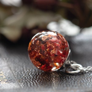 May include: A silver chain necklace with a round pendant containing dried red flowers and gold flakes. The pendant is on a dark brown surface.