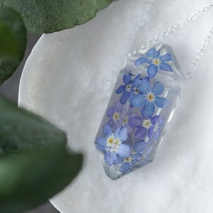 May include: A clear crystal pendant with a silver chain, featuring pressed forget-me-not flowers. The pendant is on a white surface with green leaves in the background.