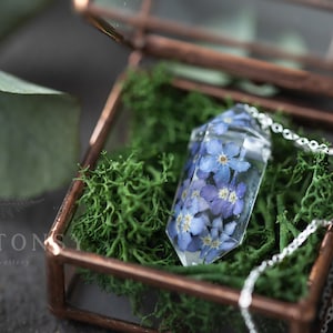 May include: A delicate silver chain necklace with a clear crystal pendant containing pressed blue forget-me-not flowers. The necklace is displayed in a small glass box with a copper frame and green moss.