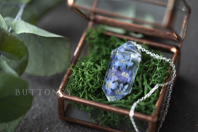 May include: A delicate silver chain necklace with a clear crystal pendant containing pressed blue forget-me-not flowers. The necklace is displayed in a small glass box with a copper frame and green moss.