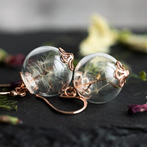 Pode incluir: Um par de brincos de ouro rosa com esferas de vidro transparente que cont&ecirc;m sementes de dente-de-le&atilde;o secas.