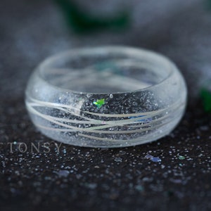 May include: A clear resin ring with silver glitter and white lines. The ring is on a dark grey surface with silver glitter.