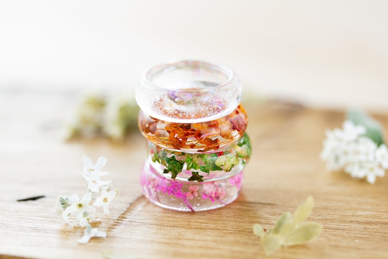 May include: Stack of four clear resin rings with embedded pressed flowers. The rings are stacked in order of colour: clear, orange, green, and pink.