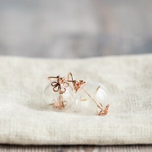 Pode incluir: Um par de brincos de ouro delicados com esferas de vidro transparentes e um delicado design floral. Os brincos est&atilde;o sobre uma superf&iacute;cie de tecido branco.