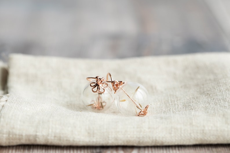 Pode incluir: Um par de brincos de ouro delicados com esferas de vidro transparentes e um delicado design floral. Os brincos est&atilde;o sobre uma superf&iacute;cie de tecido branco.