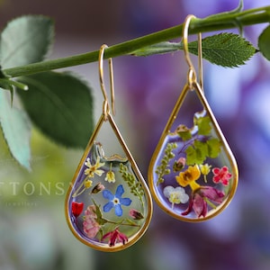 Puede incluir: Un par de pendientes de gota de oro con flores y hojas prensadas encerradas en resina transparente. Los pendientes cuelgan de una rama verde.