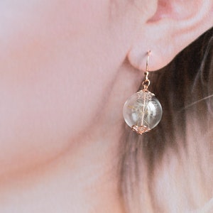 Pode incluir: Brincos de argola dourados com uma esfera de vidro transparente que cont&eacute;m um dente-de-le&atilde;o seco. A esfera est&aacute; presa a uma delicada corrente dourada.