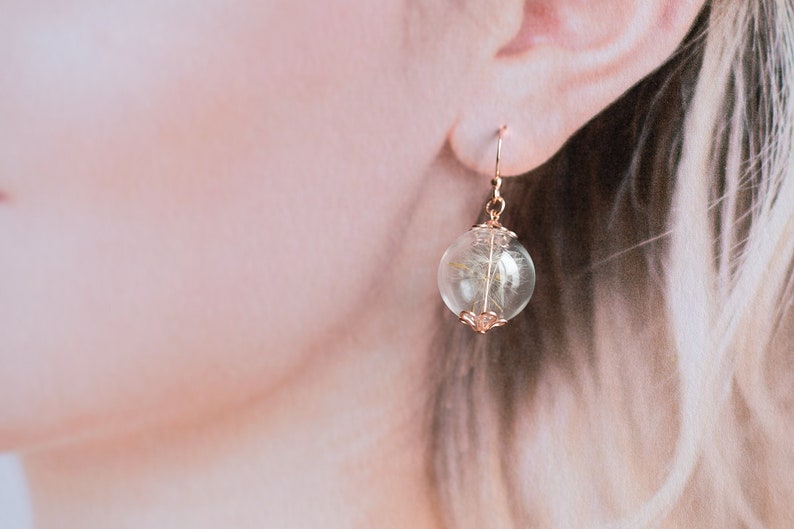 Pode incluir: Brincos de argola dourados com uma esfera de vidro transparente que cont&eacute;m um dente-de-le&atilde;o seco. A esfera est&aacute; presa a uma delicada corrente dourada.