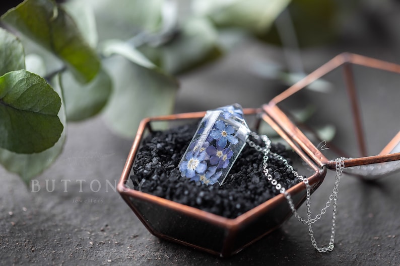 May include: A silver chain necklace with a clear glass pendant containing pressed blue forget-me-not flowers. The necklace is displayed in a hexagonal glass box with a black sand base.