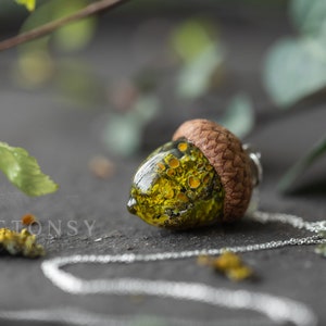 May include: A green and yellow acorn pendant necklace with a silver chain. The acorn is filled with a resin-like substance that contains small yellow and green objects.