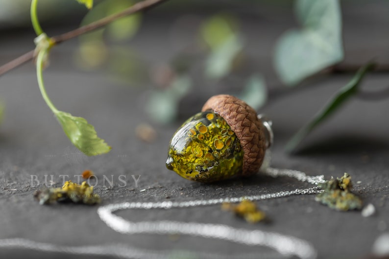 May include: A green and yellow acorn pendant necklace with a silver chain. The acorn is filled with a resin-like substance that contains small yellow and green objects.
