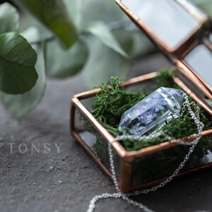 May include: A clear glass pendant with delicate blue flowers pressed inside, resting on a bed of green moss in a copper-toned glass box. The pendant is attached to a silver chain.