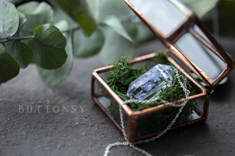 May include: A clear glass pendant with delicate blue flowers pressed inside, resting on a bed of green moss in a copper-toned glass box. The pendant is attached to a silver chain.