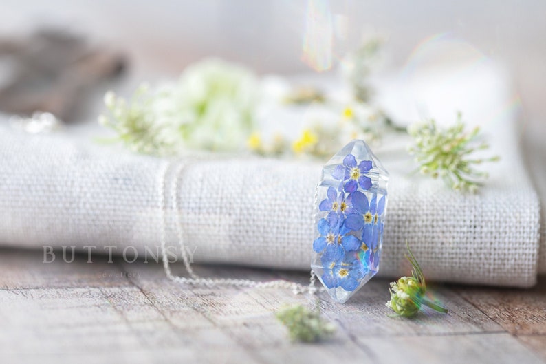 May include: A clear crystal pendant necklace with a silver chain. The pendant is filled with small blue forget-me-not flowers. The necklace is lying on a white fabric surface with white flowers.