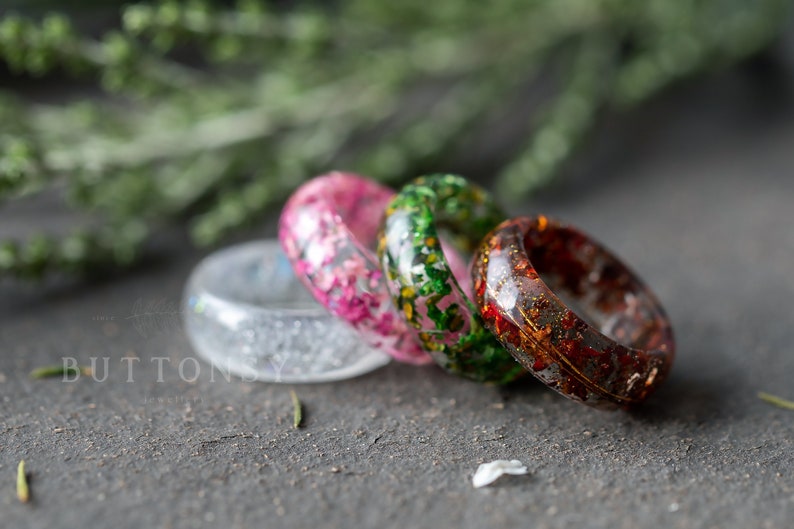 May include: Four resin rings with embedded flowers and glitter. The rings are stacked on top of each other. The rings are clear, pink, green, and orange.