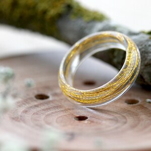 May include: A clear resin ring with gold flecks embedded throughout. The ring is sitting on a wooden surface.