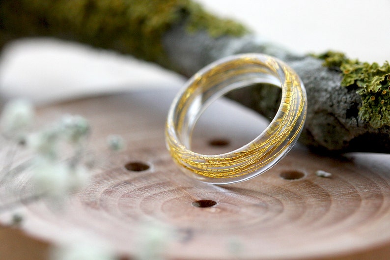 May include: A clear resin ring with gold flecks embedded throughout. The ring is sitting on a wooden surface.