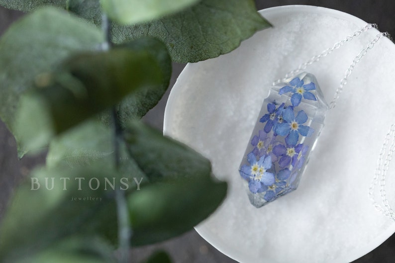 May include: A clear crystal pendant with a silver chain, featuring pressed forget-me-not flowers. The pendant is on a white surface with green leaves in the background.