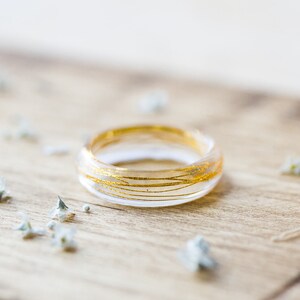 May include: A clear resin ring with gold wire embedded inside. The ring is sitting on a wooden surface with small white flowers scattered around it.