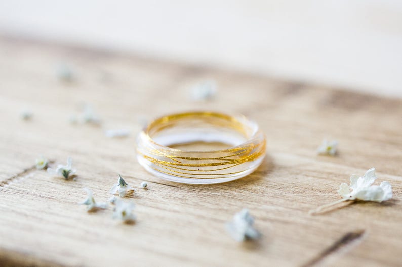 May include: A clear resin ring with gold wire embedded inside. The ring is sitting on a wooden surface with small white flowers scattered around it.