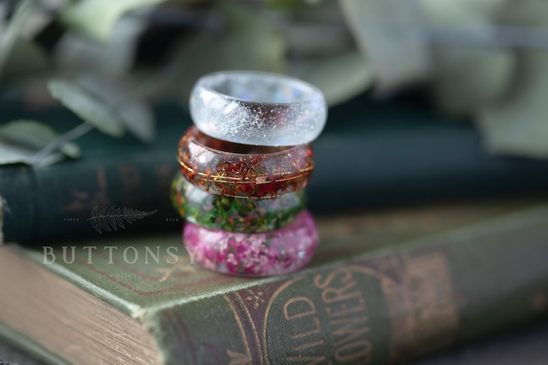 May include: Stack of four resin rings with embedded flowers and glitter. The top ring is clear with glitter, the second ring is red with gold flecks, the third ring is green with gold flecks, and the bottom ring is pink with gold flecks.