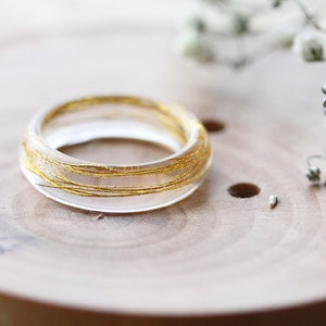 May include: A clear resin ring with gold wire embedded inside. The ring is sitting on a wooden surface.