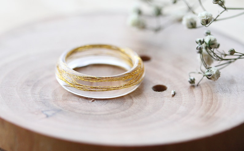May include: A clear resin ring with gold wire embedded inside. The ring is sitting on a wooden surface.