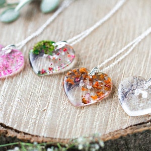 May include: Four silver necklaces with heart-shaped pendants filled with pressed flowers. The pendants are clear and show the flowers in detail. The flowers are pink, green, orange, and white.