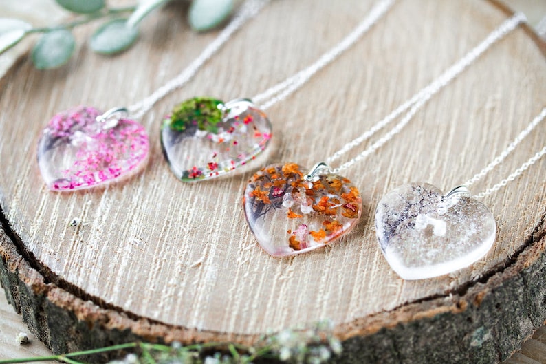May include: Four silver necklaces with heart-shaped pendants filled with pressed flowers. The pendants are clear and show the flowers in detail. The flowers are pink, green, orange, and white.