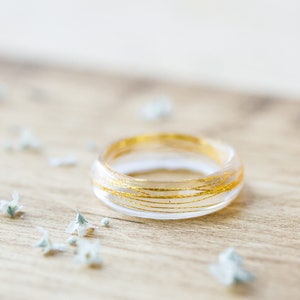 May include: A clear resin ring with thin gold wire wrapped around it. The ring is on a wooden surface.