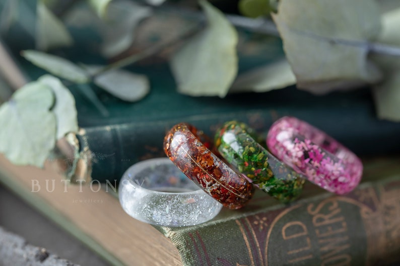 May include: Four resin rings with embedded flowers and glitter. The rings are in shades of white, green, red, and pink. The rings are arranged on a book with the word "WILD" visible.