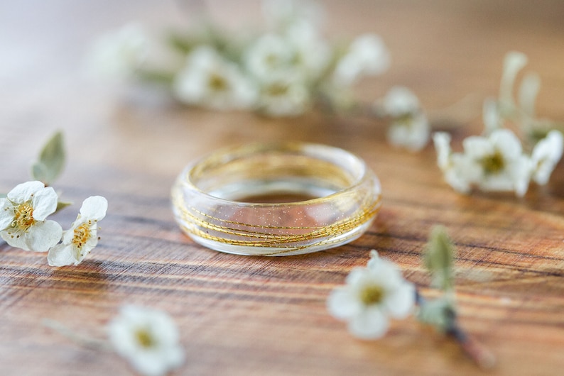 May include: A clear resin ring with gold wire embedded inside. The ring is sitting on a wooden surface with white flowers scattered around it.