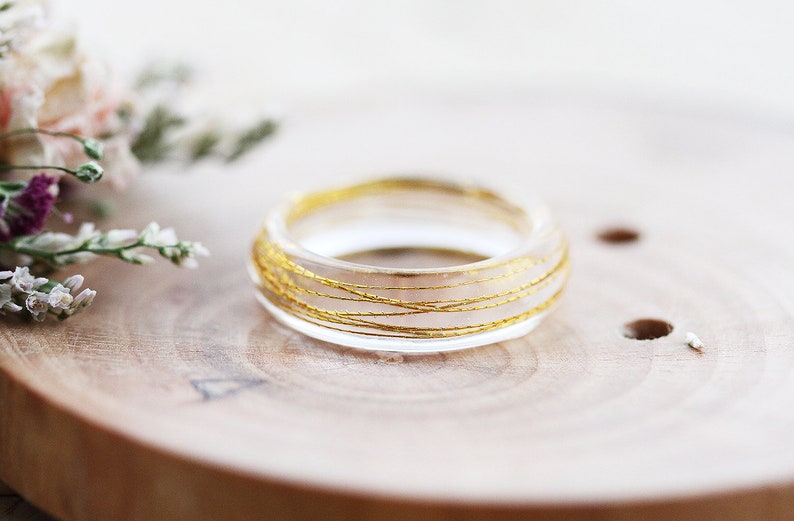May include: A clear resin ring with gold wire embedded inside. The ring is sitting on a wooden surface.