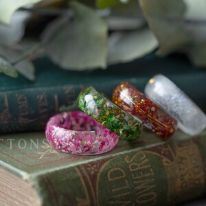 May include: Four resin rings with different pressed flower designs. The rings are stacked on top of each other on a green book with the word "BUTTONS" printed on the cover.