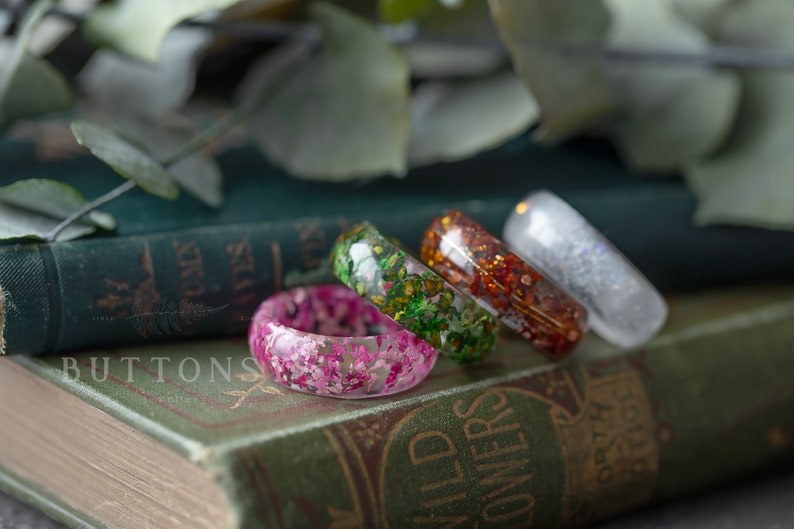 May include: Four resin rings with different pressed flower designs. The rings are stacked on top of each other on a green book with the word "BUTTONS" printed on the cover.