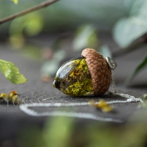 May include: A delicate necklace featuring a resin acorn pendant with a small, dried flower inside. The acorn is a natural brown color and the flower is a vibrant green.