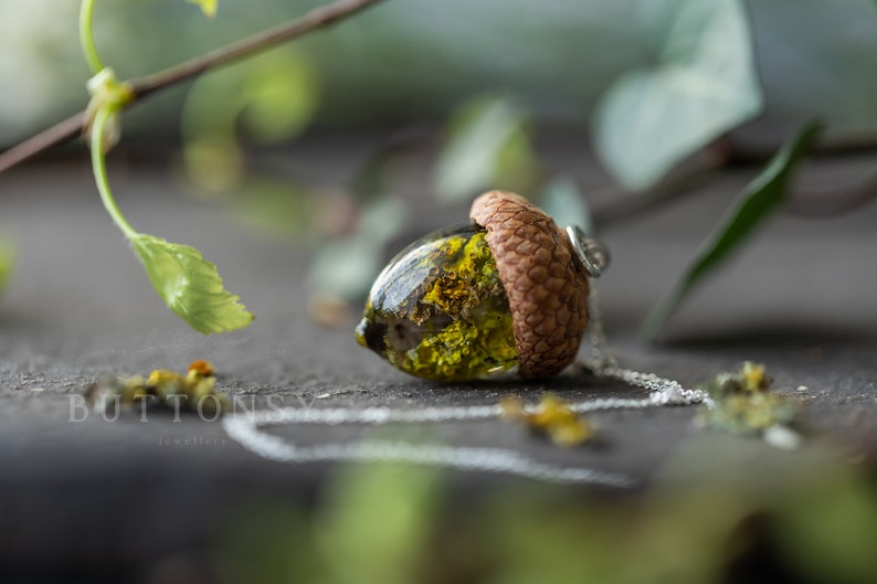 May include: A delicate necklace featuring a resin acorn pendant with a small, dried flower inside. The acorn is a natural brown color and the flower is a vibrant green.
