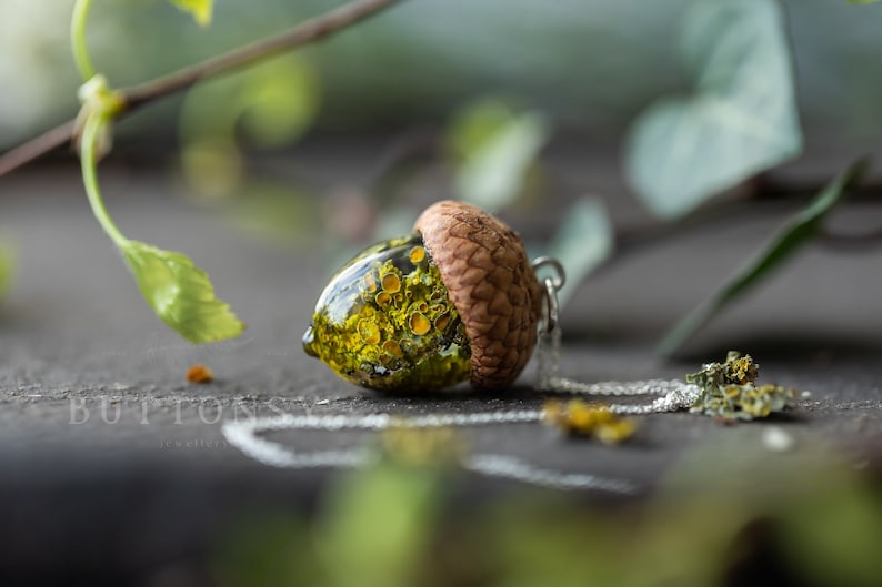 May include: A necklace with a pendant shaped like an acorn. The acorn is filled with preserved yellow and green flowers and encased in clear resin. The pendant is attached to a silver chain.