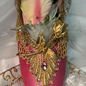 May include: A decorative pink ballet slipper adorned with gold filigree, purple gems, and a gold floral accent. The slipper is filled with pink and white roses and delicate white tulle.