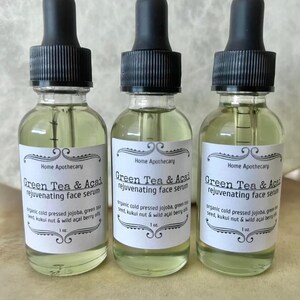 May include: Three 1-ounce bottles of green tea and acai rejuvenating face serum. The bottles are clear glass with black dropper tops and white labels. The labels have black text that reads "Home Apothecary", "Green Tea & Acai rejuvenating face serum", and "organic cold pressed jojoba, green tea seed, kukui nut & wild acai berry oils".