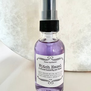 May include: A clear glass bottle with a black spray top filled with a purple liquid. The bottle has a white label with black text that reads "Home Apothecary Witch Hazel & Infused butterfly pea flower organic external use only use for healing bug bites, acne, burns, blisters, rashes, scalp irritation allergic reactions 2 oz."