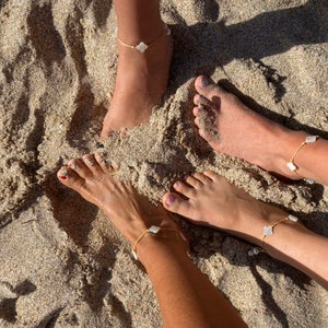 May include: Three bare feet with gold anklets adorned with white shell charms are shown on a sandy beach.