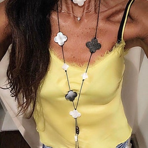 May include: A long black necklace with multiple white and black clover-shaped pendants. The necklace is layered over a thin gold chain with a small star pendant. The necklace is worn by a person wearing a yellow tank top and blue jeans.