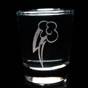 May include: A clear glass shot glass with an etched design of a stylized rainbow and cloud. The design is in white and is visible through the glass.