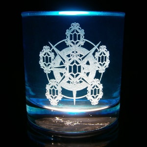 May include: A blue glass tumbler with a white etched design of a starburst with geometric shapes. The glass is illuminated from below, making the design glow.