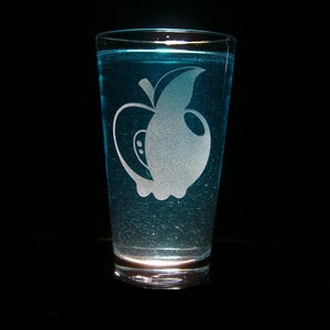 Op de afbeelding: Een helder glas met een blauwe vloeistof en een gegraveerd ontwerp van een appel. De appel is wit en heeft een steel en een blad.
