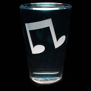 May include: A clear glass with a white musical note etched on the side, filled with a blue liquid. The glass is sitting on a black surface.