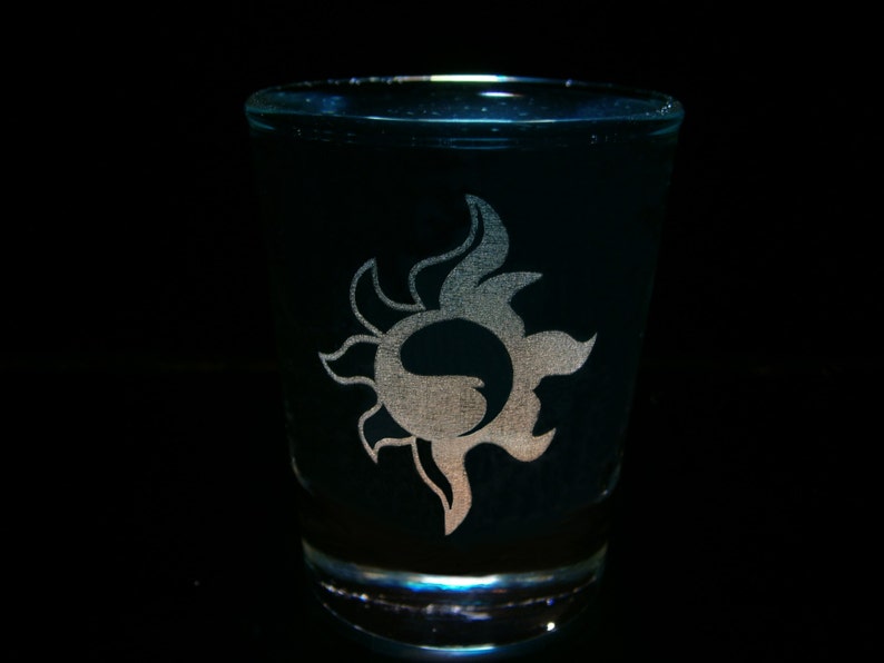May include: A dark shot glass featuring a silver sun motif. The glass is cylindrical and made of dark, possibly tinted, glass. The sun design is etched or printed on the glass, creating a contrasting effect.